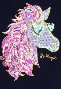 Graphisme de licorne avec des sequins roses et violets, portant des lunettes en forme de cœur. Accents dorés et le texte "Soyez magique" en script doré. Fond sombre.