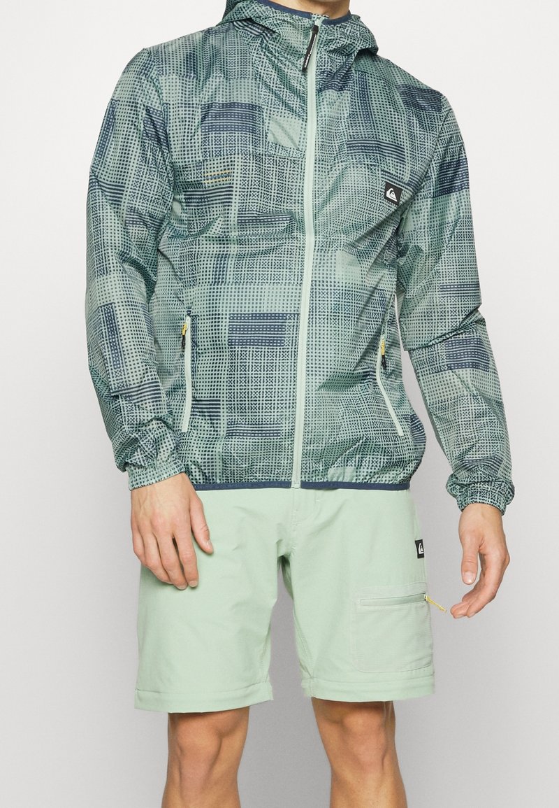 Homme portant une veste à capuche zippée verte à motifs et un short vert clair avec des poches et des détails à fermeture éclair.