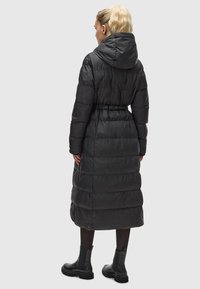 Lange zwarte pufferjas met een capuchon, met een getailleerde taille, geribbelde textuur en een lengte tot de knieën. Gecombineerd met enkellaarsjes.