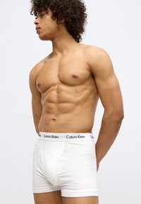 White cotton boxer briefs with a black Calvin Klein logo on the waistband, featured on a male model with curly hair, posed sideways.