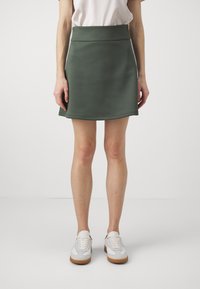 Grønn, tettsittende miniskjørt laget av glatt stoff, med bred midjebånd, flagrende hem og rene kanter. Kombinert med hvite joggesko.