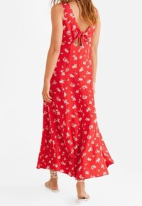 Robe maxi rouge à motifs floraux blancs, encolure dégagée et dos ouvert avec un détail noué. Tissu fluide et ourlet arrivant à la cheville.