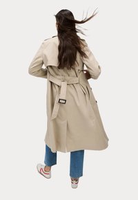 Beżowy płaszcz typu trench z paskiem w talii, dużym rozcięciem z tyłu i klasycznymi klapami. Charakteryzuje się ozdobnymi guzikami i jest zestawiony z niebieskimi dżinsami oraz białymi butami.
