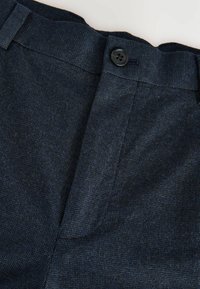 Dunkelblauer Stoff der Hose mit sichtbarem Knopf und strukturiertem Muster, das eine Nahaufnahme des Taillenbundes und des Reißverschlussbereichs zeigt.