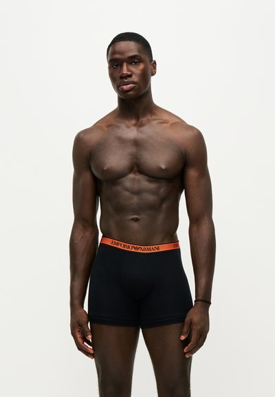 Μυώδης άνδρας που φοράει μαύρα boxer Emporio Armani με πορτοκαλί ζώνη, στέκεται μπροστά σε απλό φόντο ανοιχτού χρώματος.