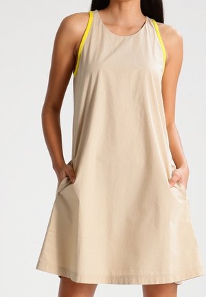 Vrouw draagt mouwloze beige A-lijn jurk met gele rand, handen in zakken, staand tegen een effen achtergrond.