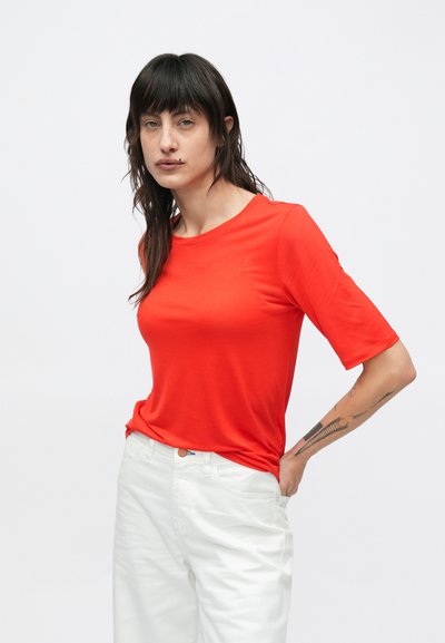 Κόκκινο κοντομάνικο μπλουζάκι με στρογγυλή λαιμόκοψη, φορεμένο με άσπρο παντελόνι. Το ύφασμα φαίνεται λείο με ελαφριά ελαστικότητα.