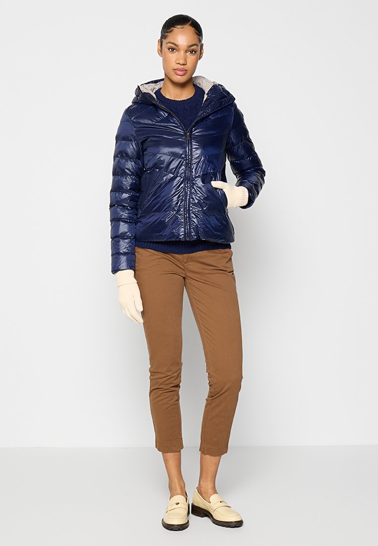 Marineblaues Steppjackett mit glänzendem Finish und Kapuze, kombiniert mit braunen Hosen. Das Modell trägt beige Handschuhe und Mokassins, was die Texturen betont.