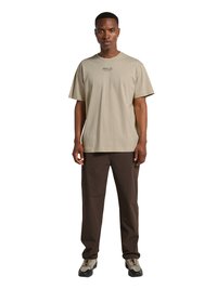 Mand iført beige HALO-brandet t-shirt, mørkebrune bukser og beige vandresko, stående med front mod kameraet på hvid baggrund.