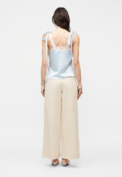 Γυναίκα με τατουάζ που φοράει ανοιχτό μπλε τοπ χωρίς μανίκια με κορδόνια στους ώμους και μπεζ παντελόνι φαρδύ, στέκεται με την πλάτη προς τα εμπρός σε απλό φόντο.