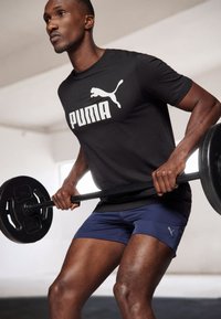 Sort t-shirt med hvid "PUMA" logo, marineblå shorts og sort vægtstænger. Glat stof, atletisk pasform, synlig muskeldefinition, minimal baggrund.