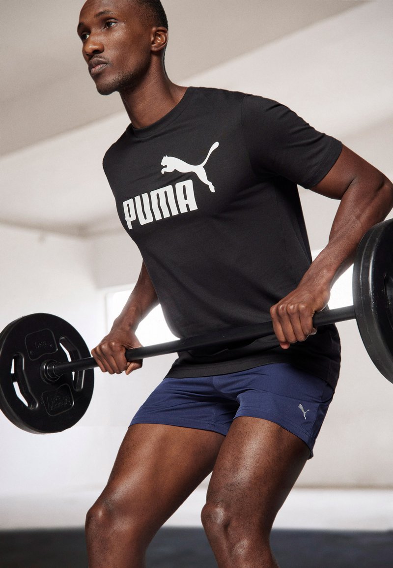 Sort t-shirt med hvid "PUMA" logo, marineblå shorts og sort vægtstænger. Glat stof, atletisk pasform, synlig muskeldefinition, minimal baggrund.