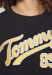 Sudadera negra con el prominente logo bordado en amarillo y blanco de "Tommy" y el número "85" en un tono plateado. Cuello redondo clásico.