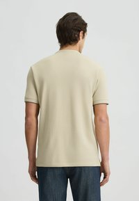 Tricou polo bej cu mânecă scurtă, cu o textură a stofei netedă, având un guler și un tiv ușor texturat, purtat cu blugi negri din denim.