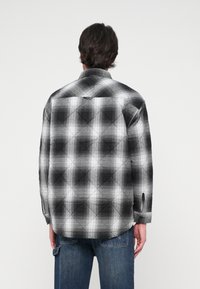 Fekete-fehér kockás ing, steppeléses textúrával, hegyes gallérral és mandzsettázott ujjakkal. Látható varrásokkal és egy hurkóval a hátulján.