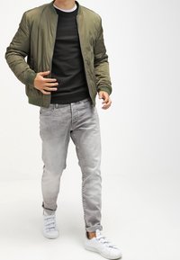 Chaqueta bomber verde oliva con puños acanalados, suéter negro, jeans gris claro y zapatillas blancas. Exterior liso y brillante en la chaqueta.