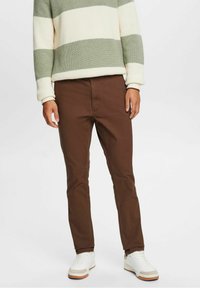 Uomo che indossa pantaloni marroni slim-fit, sneakers bianche e un maglione a righe verdi e bianche, in piedi su uno sfondo semplice.