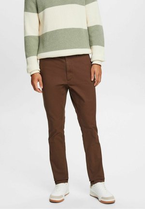 Uomo che indossa pantaloni marroni slim-fit, sneakers bianche e un maglione a righe verdi e bianche, in piedi su uno sfondo semplice.