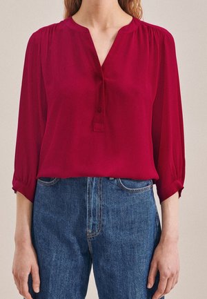 Kvinde iført en langærmet dyb rød bluse med V-hals og knapmanchetter, kombineret med højtaljede blå denimjeans.