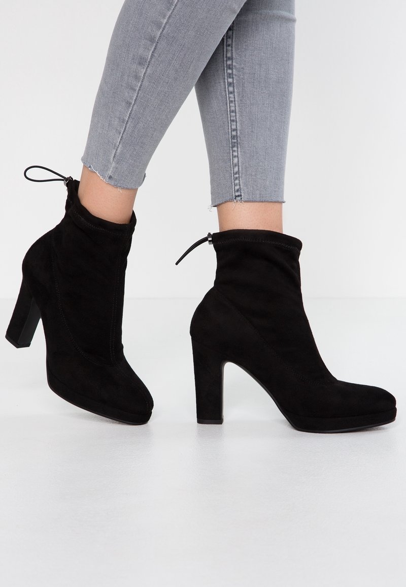 Bottines en daim noir avec un talon bloc haut, un bout arrondi et un détail à cordon coulissant au sommet. Texture lisse avec des coutures minimales.