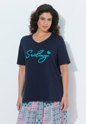 Kvinde med mørkt, krøllet hår iført en marineblå T-shirt med teksten "Smiling" og et mønstret skørt, stående op ad en hvid væg.