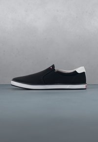 Baskets noires sans lacets avec semelle blanche et accent au talon, petit logo près des panneaux latéraux élastiques, présentées sur une surface grise avec un fond gris.