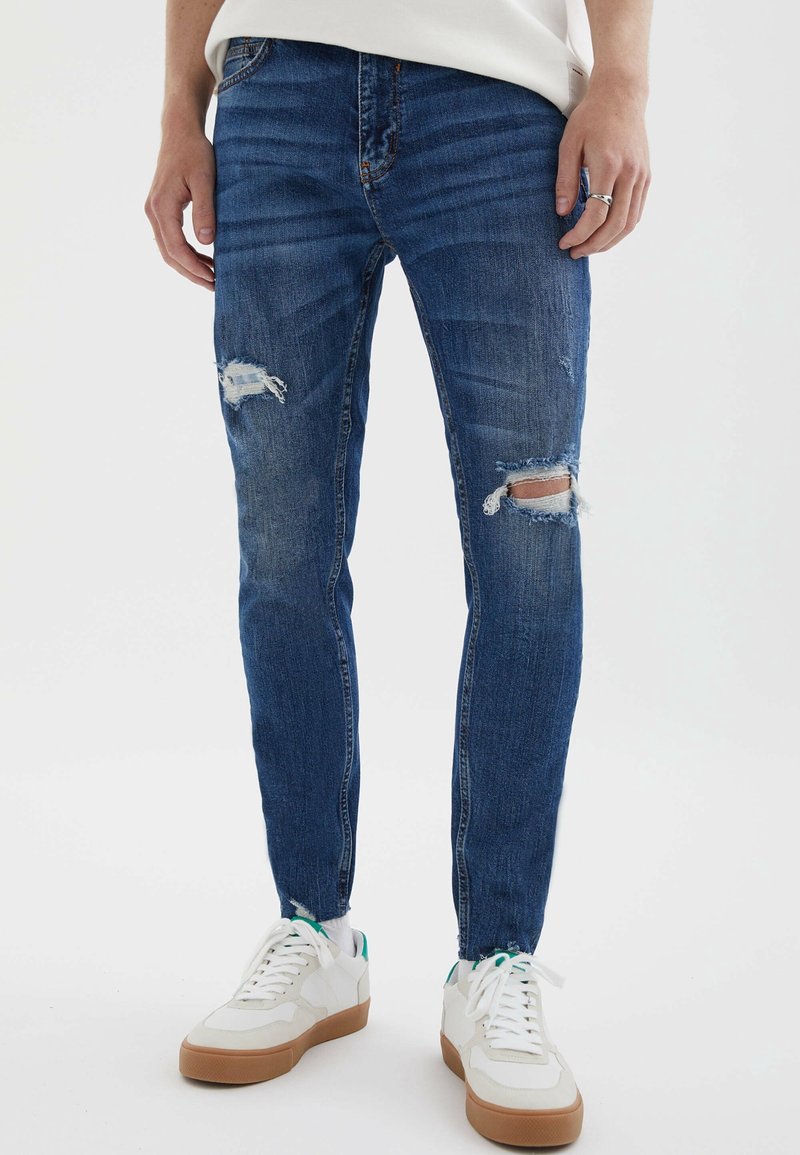 Personne portant un jean bleu slim déchiré avec des baskets blanches sur un fond blanc, mains reposant sur les côtés.