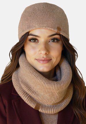 Vrouw met lang bruin haar, een beige gebreide muts en bijpassende dikke geribbelde gebreide nekwarmer, gedragen over een kastanjebruine jas.