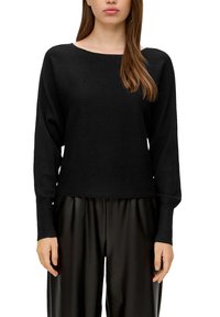 Schwarzer gerippter Pullover mit einem Bootsausschnitt und langen Ärmeln, ausgestattet mit einem lockeren Schnitt und strukturiertem Strick, kombiniert mit schwarzen plissierten Hosen.