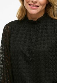 Blusa nera in pizzo con motivi floreali testurizzati, colletto alto e maniche lunghe, con un design trasparente e bordi a scallop.