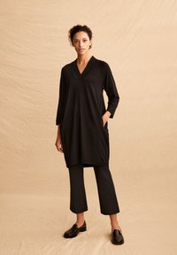 Schwarzes Tunika mit V-Ausschnitt, Dreiviertelärmeln und seitlichen Taschen; kombiniert mit schwarzen, knöchellangen Hosen und schwarzen Schlupfschuhen.