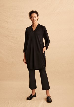 Schwarzes Tunika mit V-Ausschnitt, Dreiviertelärmeln und seitlichen Taschen; kombiniert mit schwarzen, knöchellangen Hosen und schwarzen Schlupfschuhen.