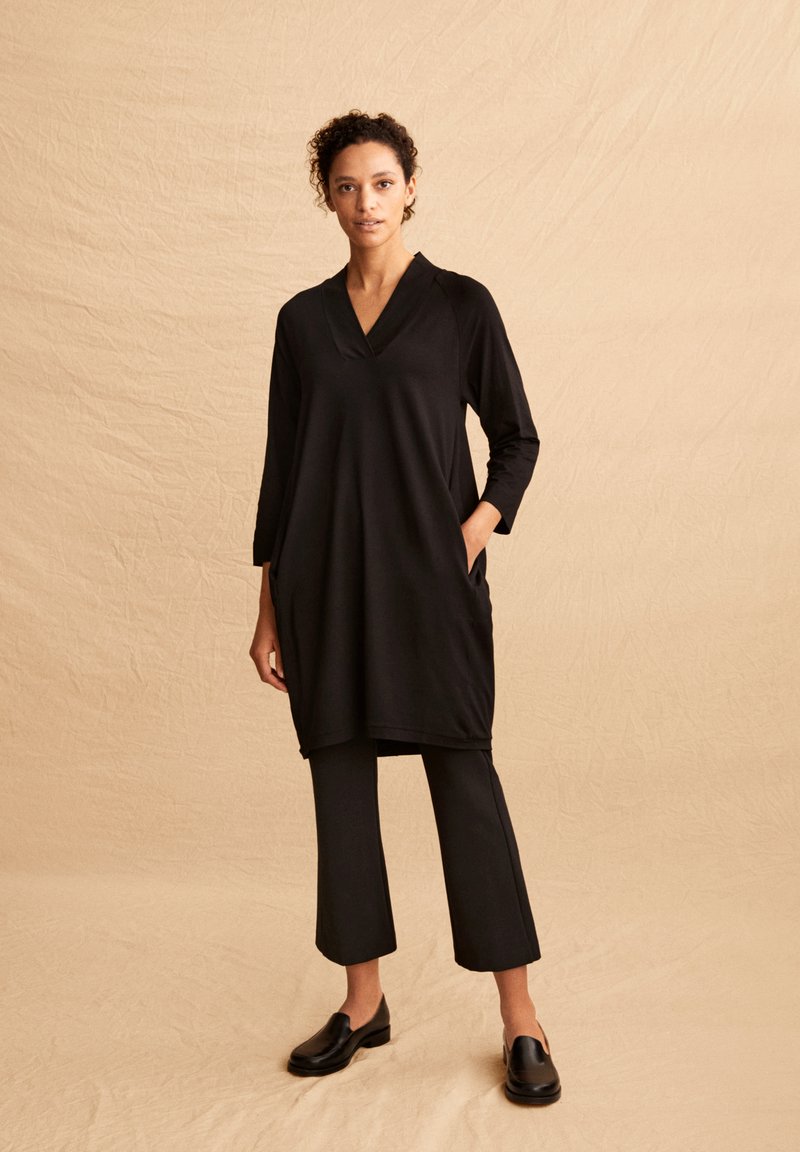 Schwarzes Tunika mit V-Ausschnitt, Dreiviertelärmeln und seitlichen Taschen; kombiniert mit schwarzen, knöchellangen Hosen und schwarzen Schlupfschuhen.