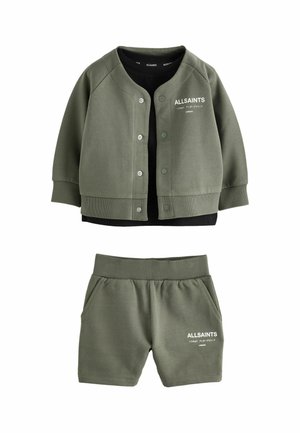 Jeune garçon debout portant une veste et un short vert olive AllSaints, un t-shirt noir, des chaussettes blanches et des baskets blanches sur fond clair.