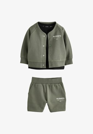 Ung dreng stående iført olivengrøn AllSaints jakke og shorts, sort skjorte, hvide sokker og hvide sneakers på lys baggrund.
