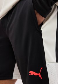 Μαύρα αθλητικά σορτς με κόκκινο λογότυπο Puma και λευκές πλαϊνές πινελιές, φορεμένα με το χέρι εν μέρει μέσα στην τσέπη.