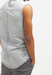 Maglietta senza maniche grigia chiara con una texture sottile, orlo curvo e due toppe decorate sul lato inferiore, indossata sopra pantaloni grigi.