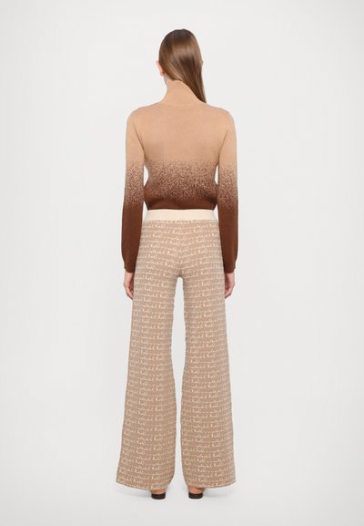 Pleteni pulover s turtleneck iz gradientnih odtenkov bež do rjave, v kombinaciji s širokimi hlačami, ki imajo ponavljajoč se tekstovni vzorec v krem barvi.