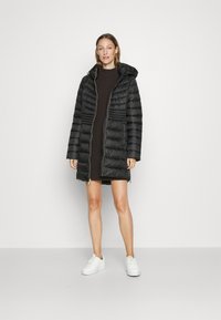 Zwarte pufferjas met capuchon, rits aan de voorkant en ribbelstructuur. Bevat zijzakken en een knielange stijl, gecombineerd met witte sneakers.
