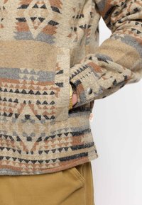 Strickpullover mit strukturiertem Muster in Beige, Braun und Grau. Mit einer Fronttasche und geknöpften Bündchen mit einem Label.