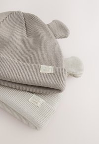 Kaksi neulottua vauvan pipoa: yksi vaaleanharmaa ja yksi luonnonvalkoinen. Molemmissa on joustinneulosta ja päällä karhun korvat. Merkkilapussa lukee "pikkuinen".