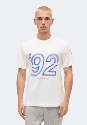 Mladić s kratkom plavom kosom nosi bijelu majicu s velikim plavim "92" i crvenim tekstom "Manchester United", u kombinaciji sa sivim hlačama.