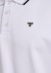 Hvid polo-shirt med tre knapper og et sort-hvidt biemblem på venstre bryst under kraven.
