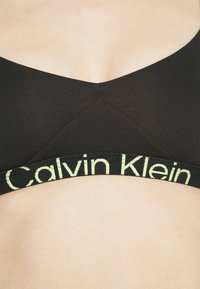 Bralette nera con design a scollo a V, texture a costine e un elastico contrastante con la scritta "Calvin Klein" in lettere verde chiaro.