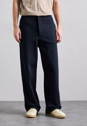 Navy loose-fit pants made of soft fabric with a front zipper and hook closure, paired with cream-colored sneakers featuring a textured design.