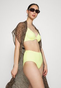 Bikini amarelo com bordas em forma de onda, combinado com um vestido transparente de estampado leopardo castanho. Modelo usando óculos de sol escuros, cabelo preso.