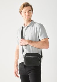 Sac bandoulière noir avec un design texturé, un compartiment zippé et une sangle ajustable, tenu par une personne portant une chemise gris clair.