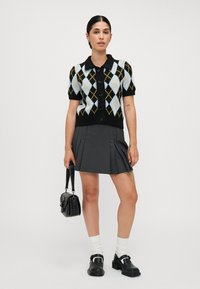 Černý krátký rukáv cardigan s argyle vzorem v modré a žluté, s límcem a knoflíky. Spárováno se šedou plisovanou sukní.