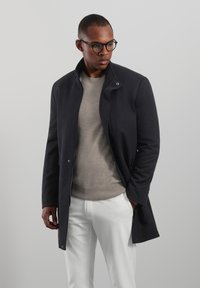 Marineblauwe jas met een hoge kraag, drukknoopsluiting aan de voorkant, zijzakken en een gladde textuur, gedragen over een lichtgrijze trui en witte broek.