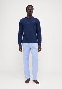 Jūras spēku garās piedurknes krekls ar pogu plāksni un sarkano "POLO" logo, apvienots ar gaiši ziliem biksēm, valkāts basām kājām uz vienkārša fona.